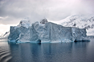 A large iceberg floating in the ocean with snow-covered mountains in the background, under a cloudy sky.